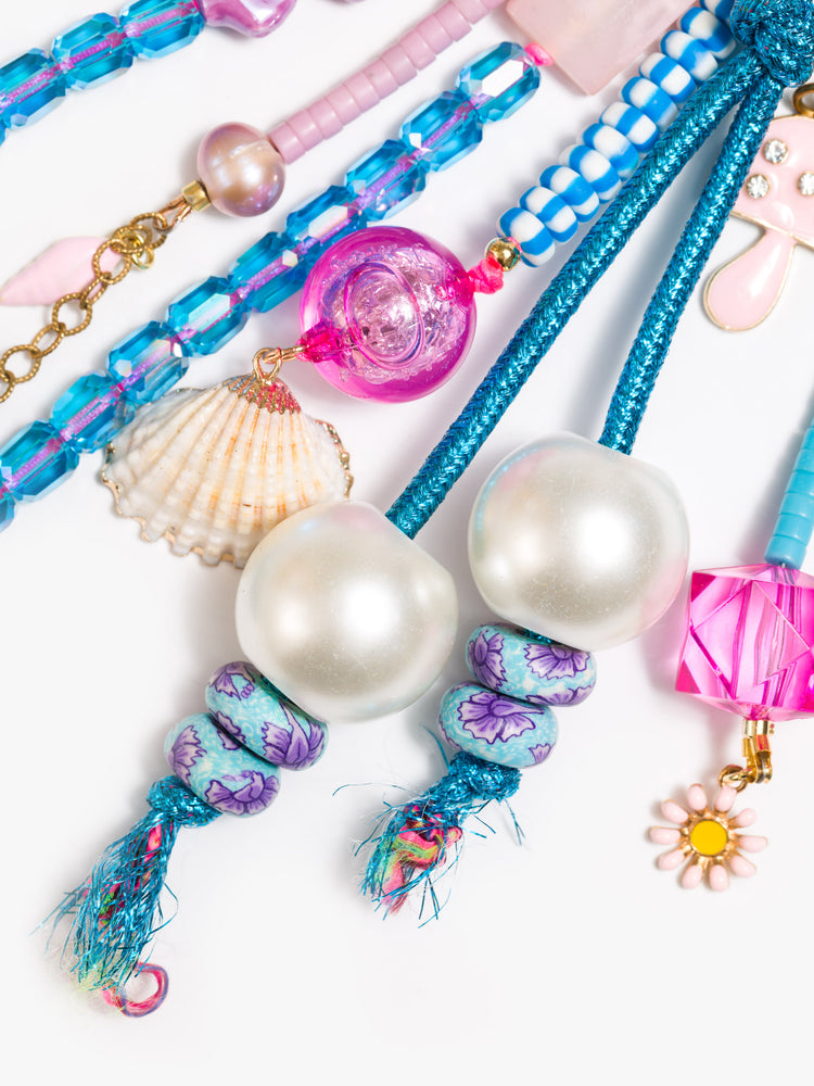 Close up view of a bag charm with mixed beads in shades of purple, blue, pink and pearl, with shell, flower and mushroom charms. 