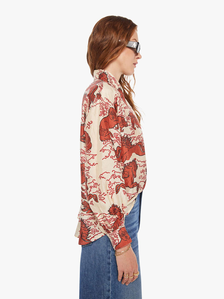Side  view of a woman in a cream shirt with a maroon Pegasus print, and features drop shoulders, long sleeves, curved hems and buttons down the front.