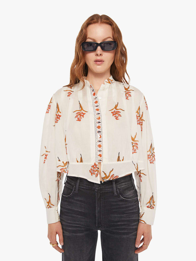 Front view of a woman in a off-white shirt with a delicate floral print, and features a crewneck, pleats along the shoulder, long balloon sleeves, a cropped fit and buttons down the front.