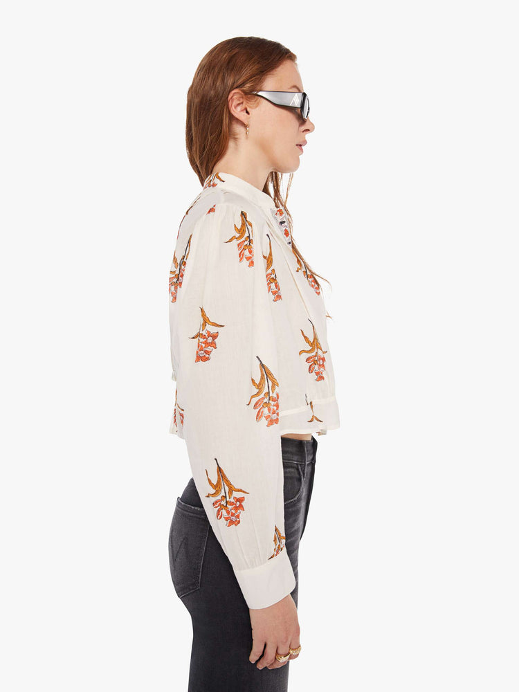 Side  view of a woman in a off-white shirt with a delicate floral print, and features a crewneck, pleats along the shoulder, long balloon sleeves, a cropped fit and buttons down the front.