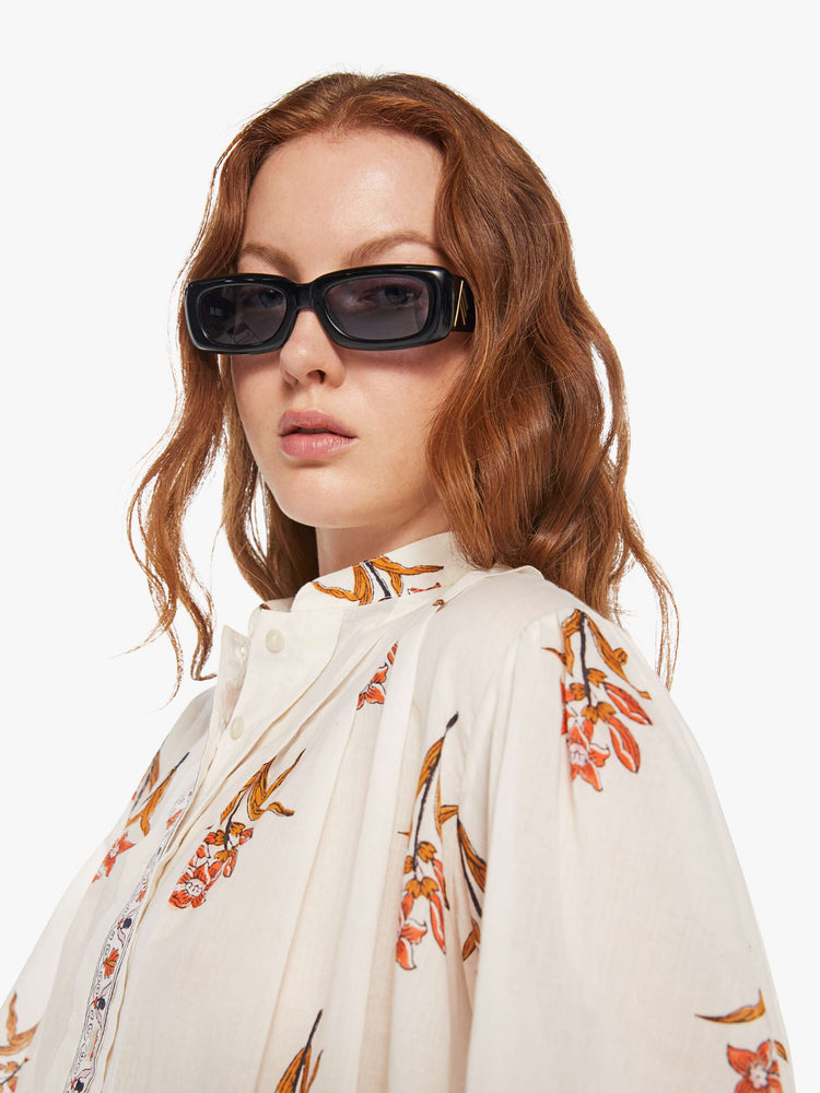 Close up  view of a woman in a off-white shirt with a delicate floral print, and features a crewneck, pleats along the shoulder, long balloon sleeves, a cropped fit and buttons down the front.