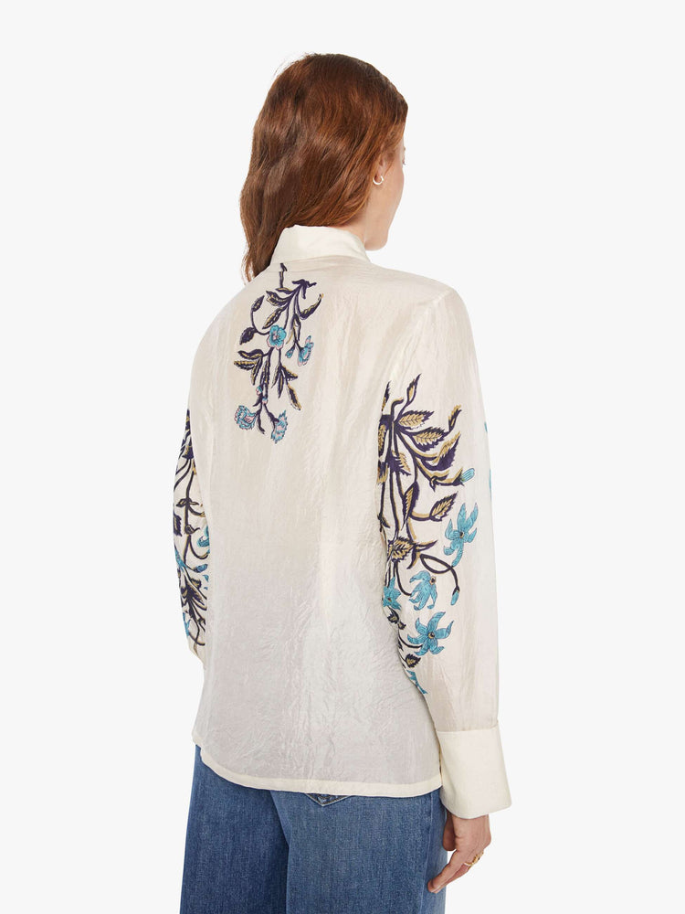 Back view of a woman in an off-white shirt with blue lilies across the chest and sleeves, and features long balloon sleeves and a loose fit.