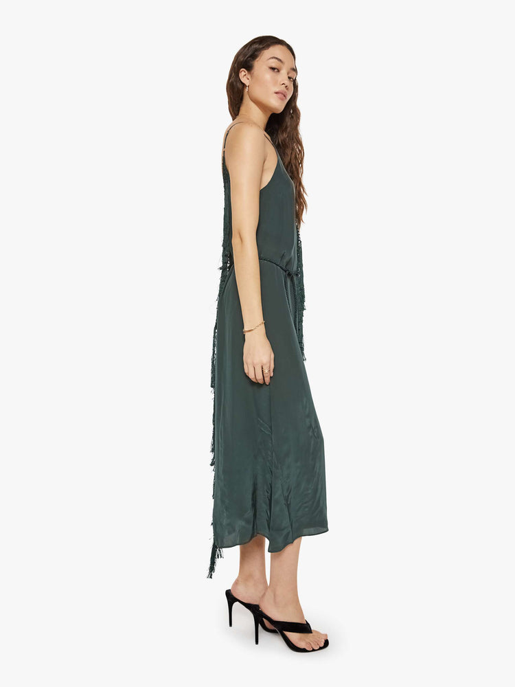 Side view of a woman in a dark green dress designed with thin spaghetti straps, a deep V-neck, fringed trim and a flowy fit.