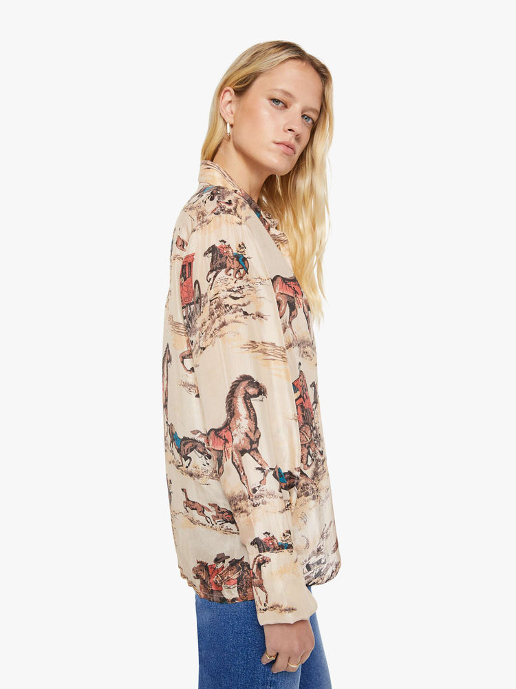 Side view of a woman in a button up peachy cream with cowboy motifs, and features long balloon sleeves and a loose fit.