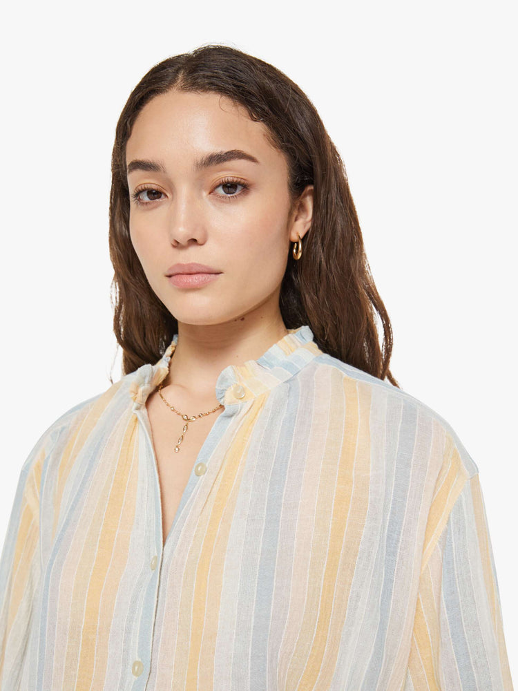 Close up view of a woman in a vertical yellow, white and light blue stripes, and features a stacked collar shirt, drop shoulders, long balloon sleeves and a curved hem.