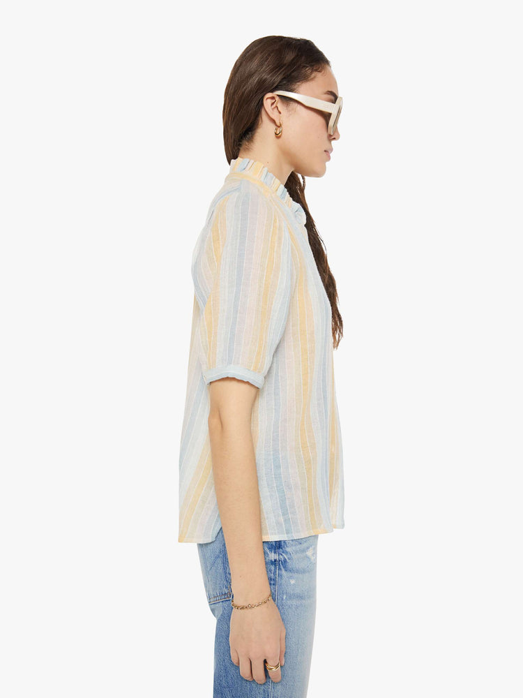 Side view of a woman in a vertical yellow, white and light blue stripes, and features a stacked collar shirt with an elbow-length sleeves, buttons down the front and a curved hem.