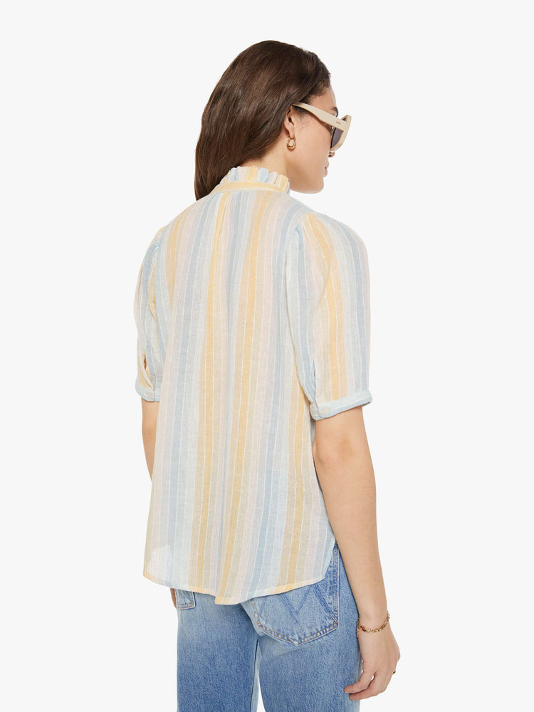 Back view of a woman in a vertical yellow, white and light blue stripes, and features a stacked collar shirt with an elbow-length sleeves, buttons down the front and a curved hem.