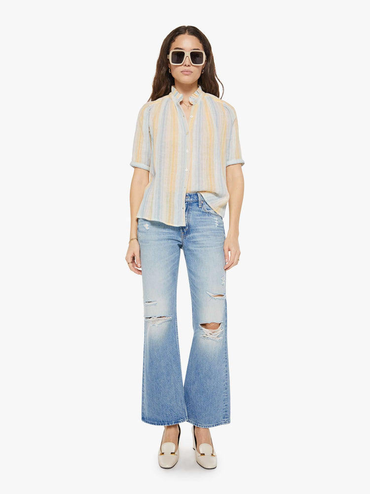 Full body view of a woman in a vertical yellow, white and light blue stripes, and features a stacked collar shirt with an elbow-length sleeves, buttons down the front and a curved hem.