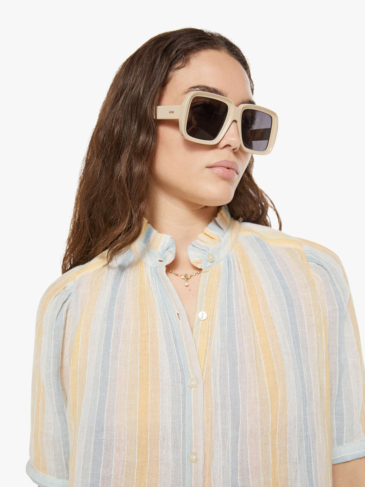 Close up view of a woman in a vertical yellow, white and light blue stripes, and features a stacked collar shirt with an elbow-length sleeves, buttons down the front and a curved hem.
