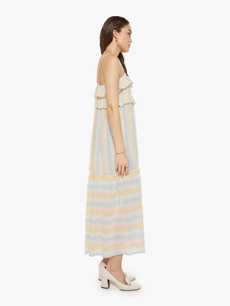 Side view of a woman in a vertical yellow, white and light blue stripes dress with thin spaghetti straps, a square neck with ruffles, a loose, tiered skirt and an ankle-length hem.