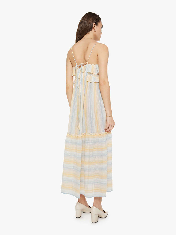 Back view of a woman in a vertical yellow, white and light blue stripes dress with thin spaghetti straps, a square neck with ruffles, a loose, tiered skirt and an ankle-length hem.