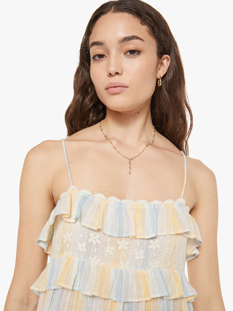 Close up view of a woman in a vertical yellow, white and light blue stripes dress with thin spaghetti straps, a square neck with ruffles, a loose, tiered skirt and an ankle-length hem.