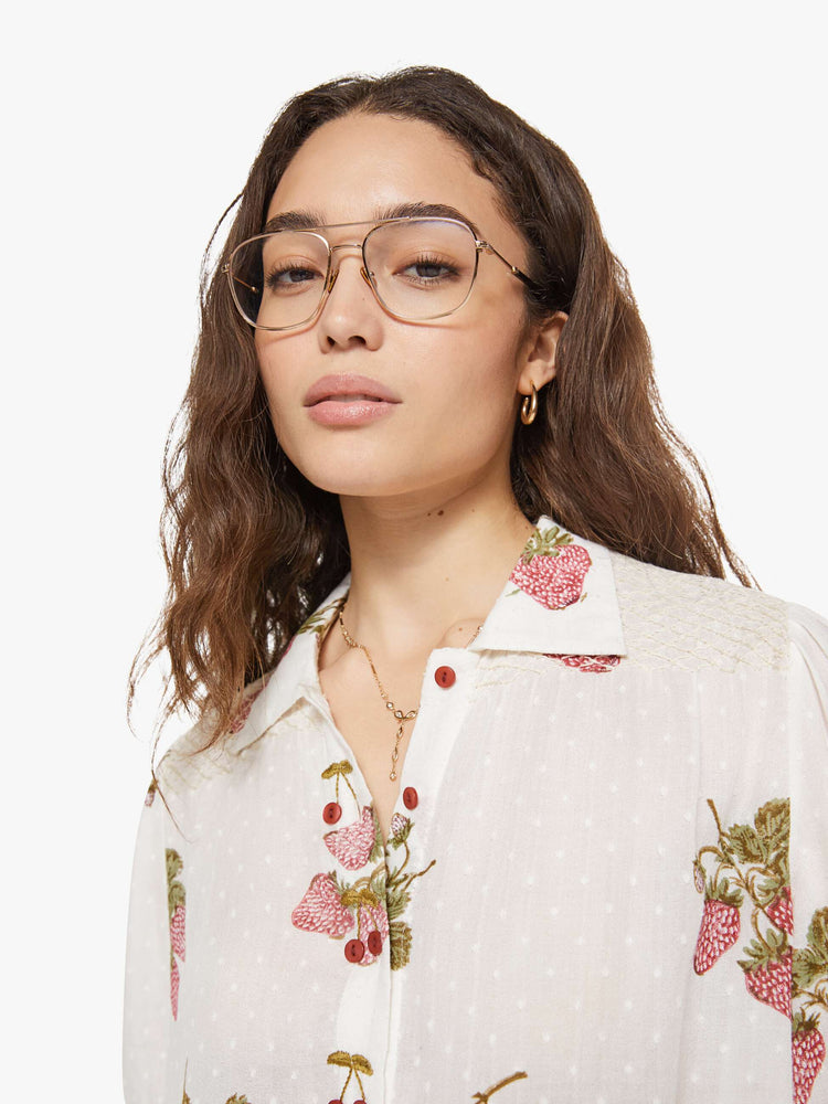 Close up view of a woman in an off-white shirt with a faded strawberry print, and is designed with a collared neck, short sleeves and buttons down the front.