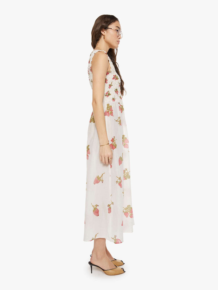 Side view of a woman in an off-white with a faded strawberry print dress with a curved square neck, wide straps, a fitted bodice and a flowy length skirt with an ankle-length hem.