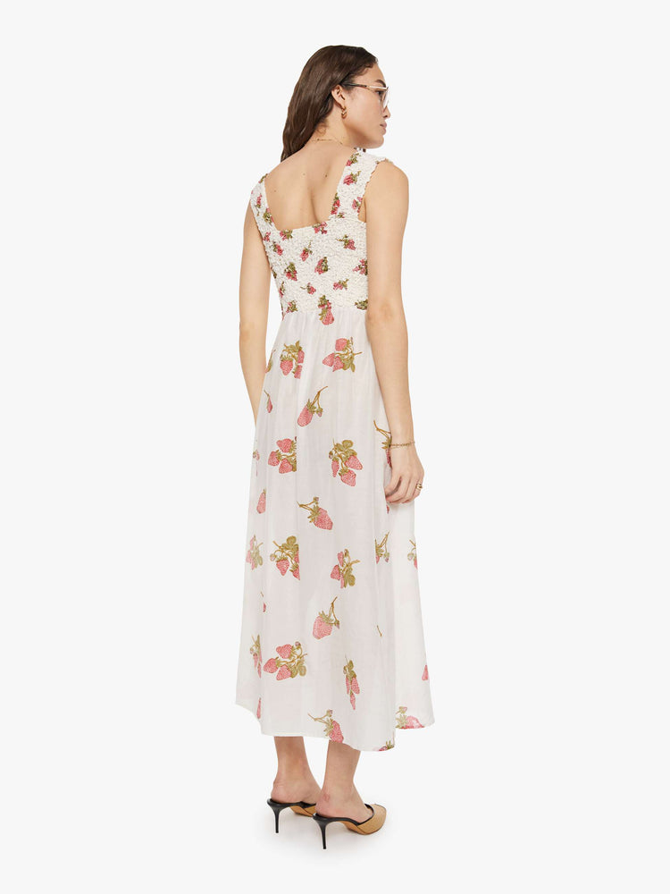 Back view of a woman in an off-white with a faded strawberry print dress with a curved square neck, wide straps, a fitted bodice and a flowy length skirt with an ankle-length hem.