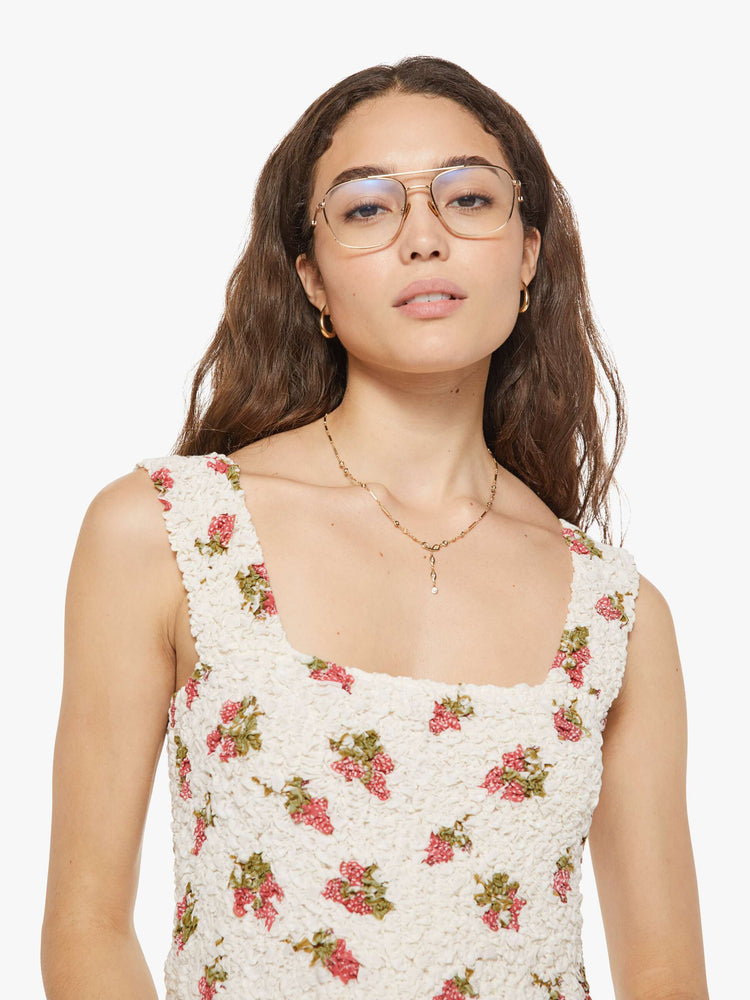 Close up view of a woman in an off-white with a faded strawberry print dress with a curved square neck, wide straps, a fitted bodice and a flowy length skirt with an ankle-length hem.