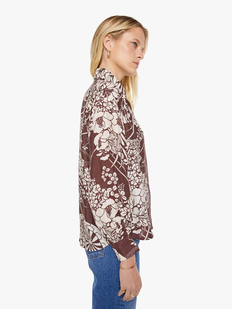 Side view of a woman in a brown with a white vintage-inspired print shirt that features drop shoulders, long sleeves, curved hems and buttons down the front.