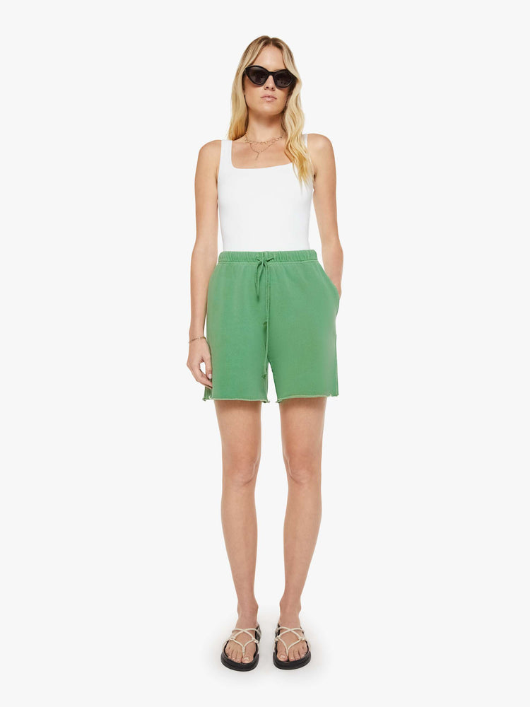 Front full image of a woman in green sweatshort is designed with a drawstring waistband, slit pockets, raw hems and a loose fit.