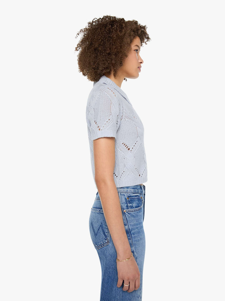 Side view of a woman in a light blue-grey top  designed with short sleeves, a collared V-neck that buttons, ribbed hems and a cable knit pattern.