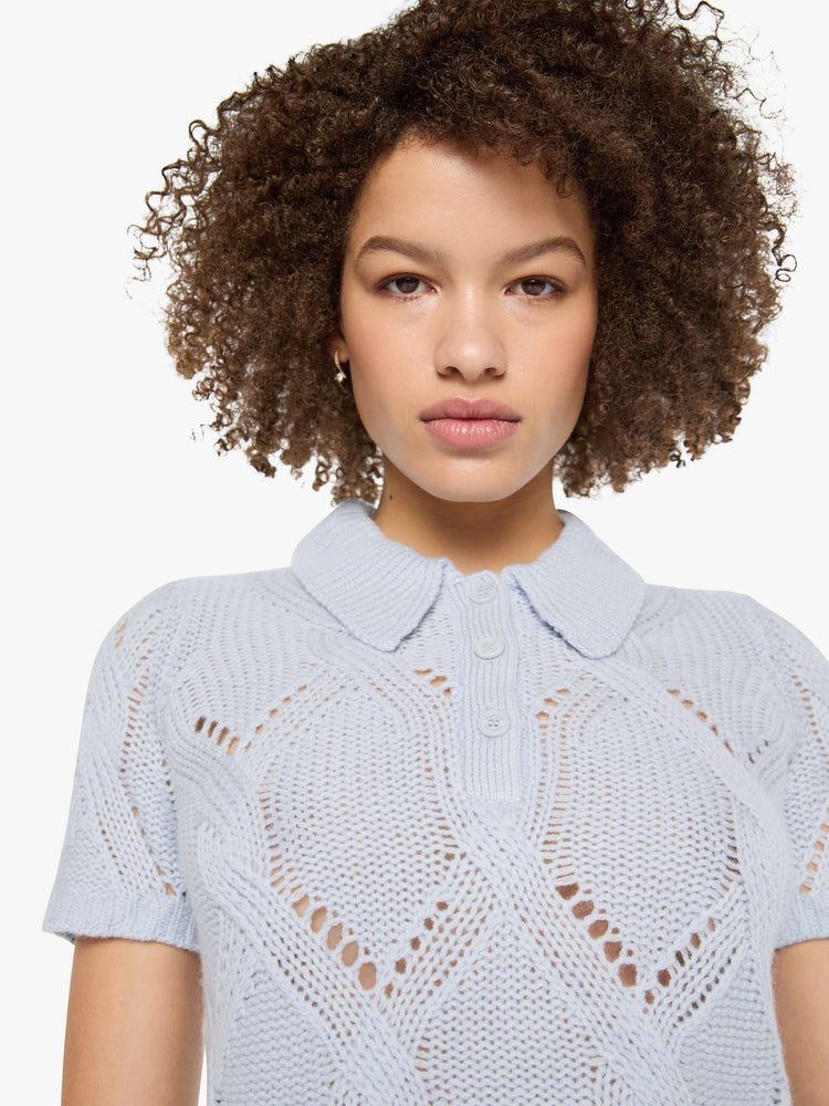 Close up view of a woman in a light blue-grey top  designed with short sleeves, a collared V-neck that buttons, ribbed hems and a cable knit pattern.