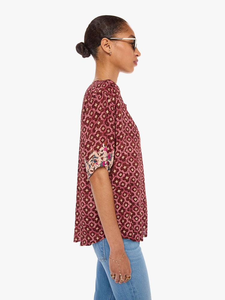 Side view of a woman in a brick red mosaic print deep V-neck, buttons down the front, short sleeves and is slightly pleated under the collar for a loose, flowy fit.