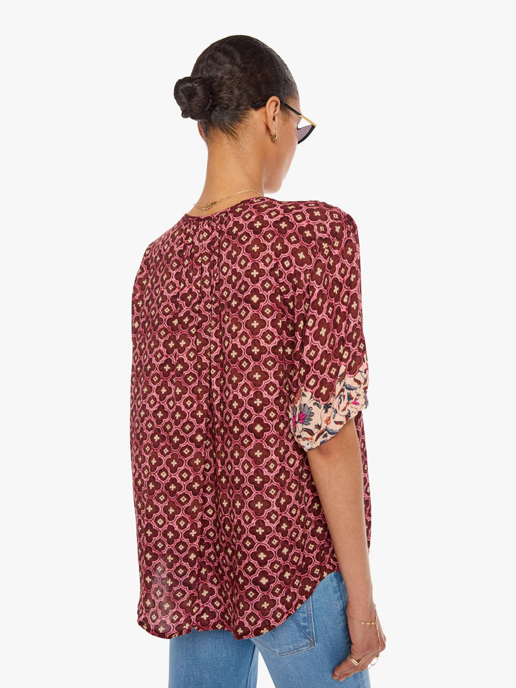 Back view of a woman in a brick red mosaic print deep V-neck, buttons down the front, short sleeves and is slightly pleated under the collar for a loose, flowy fit.