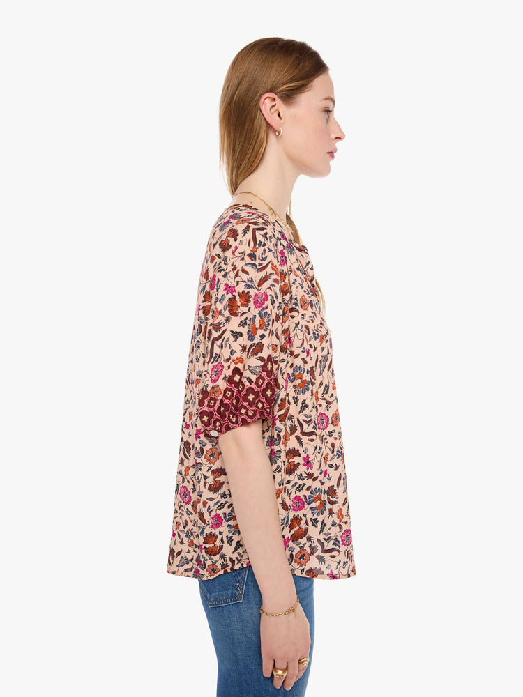 Side view of a woman in cream with a warm-toned floral print top features a deep V-neck, buttons down the front, short sleeves and is slightly pleated under the collar for a loose, flowy fit.