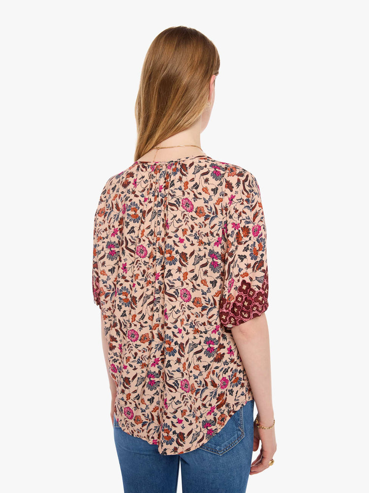 Back view of a woman in cream with a warm-toned floral print top features a deep V-neck, buttons down the front, short sleeves and is slightly pleated under the collar for a loose, flowy fit.