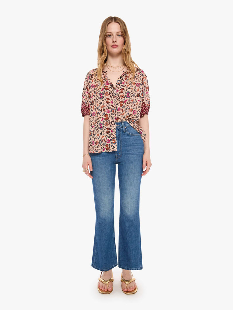 Full body view of a woman in cream with a warm-toned floral print top features a deep V-neck, buttons down the front, short sleeves and is slightly pleated under the collar for a loose, flowy fit.