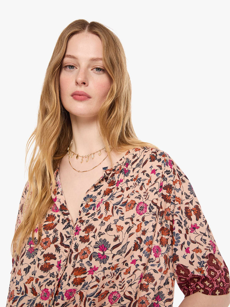 Close up view of a woman in cream with a warm-toned floral print top features a deep V-neck, buttons down the front, short sleeves and is slightly pleated under the collar for a loose, flowy fit.