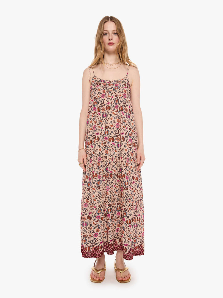 Front view of a woman in a cream with a warm-toned floral print dress features thin spaghetti straps, a scoop neck and a tiered, ankle-length skirt.