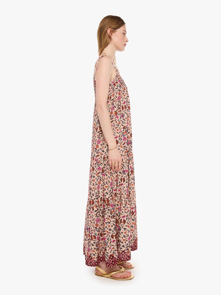 Side view of a woman in a cream with a warm-toned floral print dress features thin spaghetti straps, a scoop neck and a tiered, ankle-length skirt.