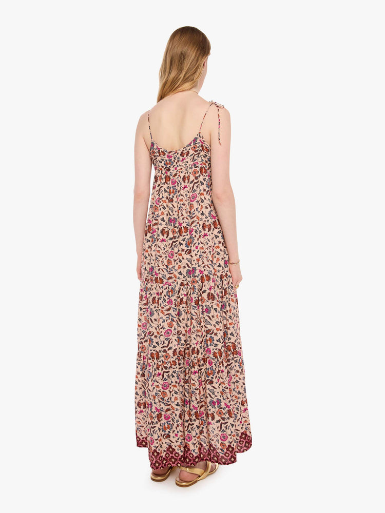 Back view of a woman in a cream with a warm-toned floral print dress features thin spaghetti straps, a scoop neck and a tiered, ankle-length skirt.