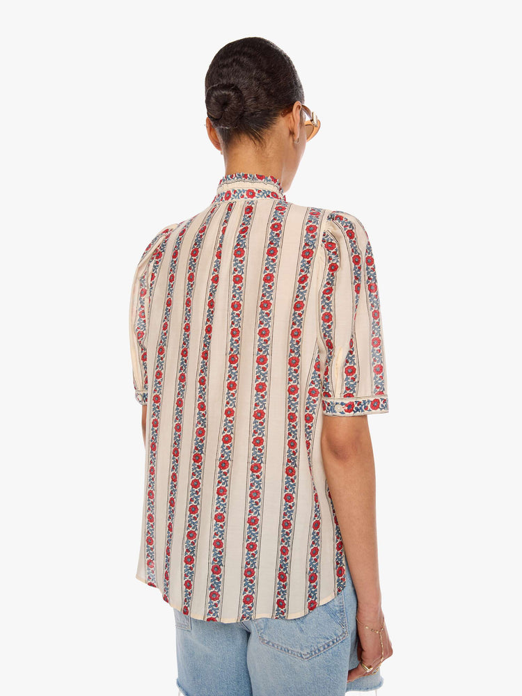 Back view of a woman in a silk cream with light blue stripes detailed with roses shirt with a stacked collar, elbow-length sleeves, buttons down the front and a curved hem.
