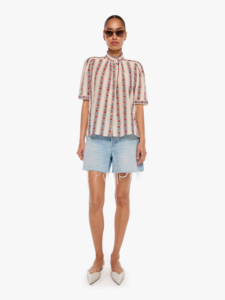 Full body view of a woman in a silk cream with light blue stripes detailed with roses shirt with a stacked collar, elbow-length sleeves, buttons down the front and a curved hem.