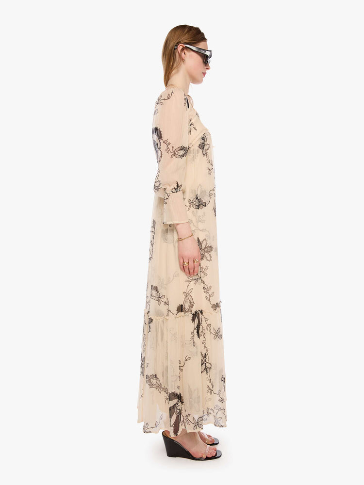 Side view of a woman in a lace printed chiffon overlay dress and features a rounded square neck, long balloon sleeves with elastic hems and a loose, flowy skirt with an ankle-length hem.