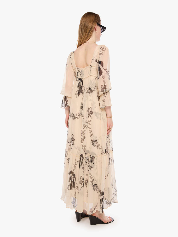 Back view of a woman in a lace printed chiffon overlay dress and features a rounded square neck, long balloon sleeves with elastic hems and a loose, flowy skirt with an ankle-length hem.