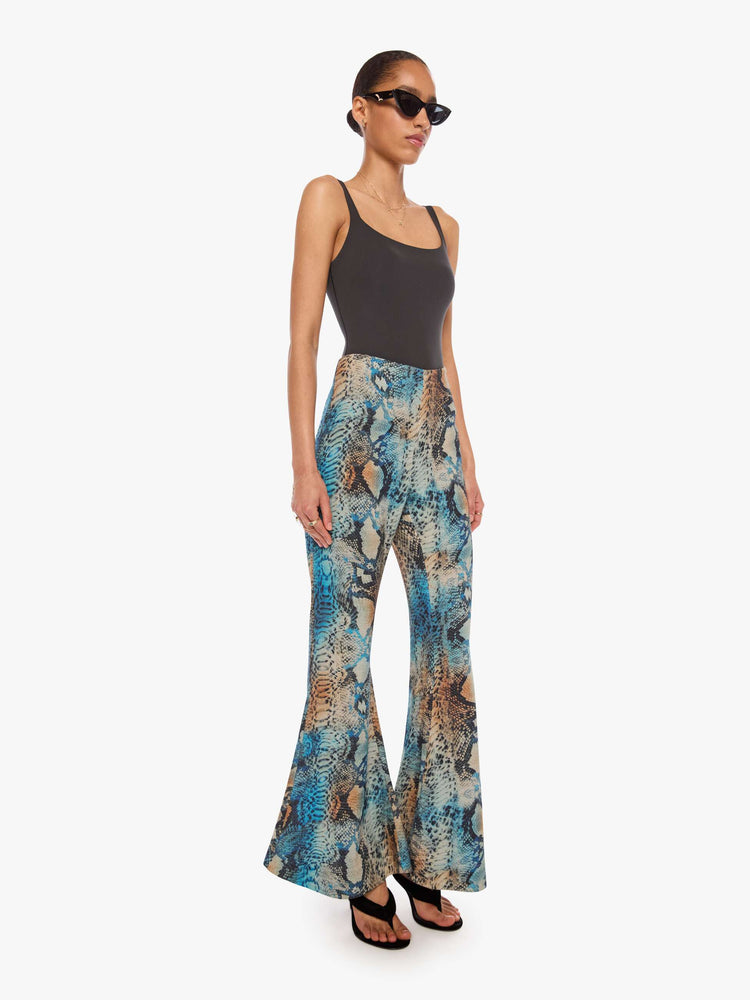 Side angle view of a woman in a pant with a blue, cream, brown and black oversized snake print, and features a high rise and a loose, flared leg.