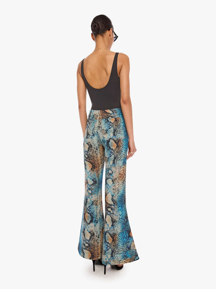 Back view of a woman in a pant with a blue, cream, brown and black oversized snake print, and features a high rise and a loose, flared leg.