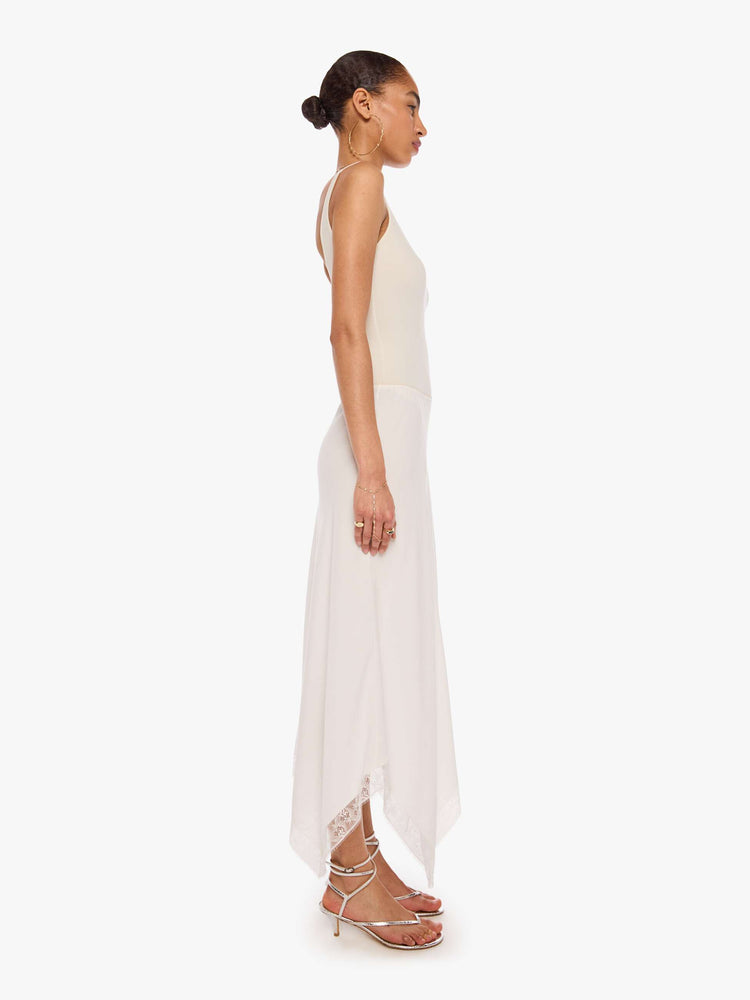 Side view of a woman in a white bias-cut maxi skirt features a flowy fit and an ankle-length uneven hem with lace trim.