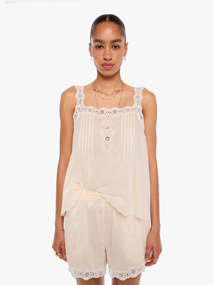 Front view of a woman in an off-white  shirt with pleats, embroidery and eyelet details on the chest and straps and a scalloped hem.