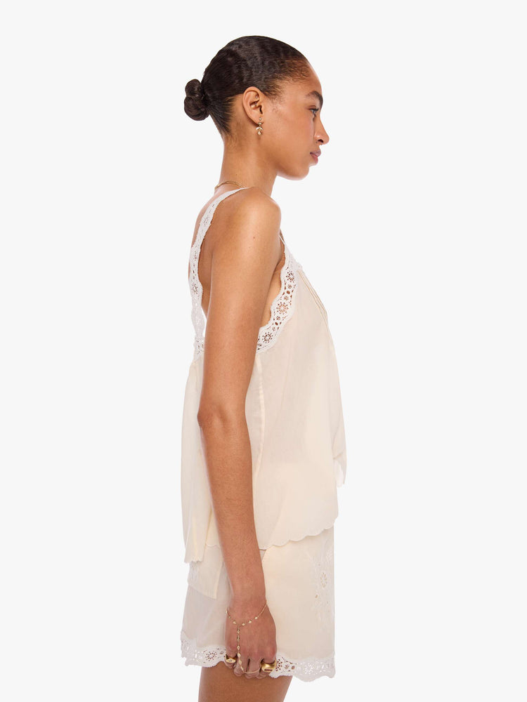 Side view of a woman in an off-white  shirt with pleats, embroidery and eyelet details on the chest and straps and a scalloped hem.