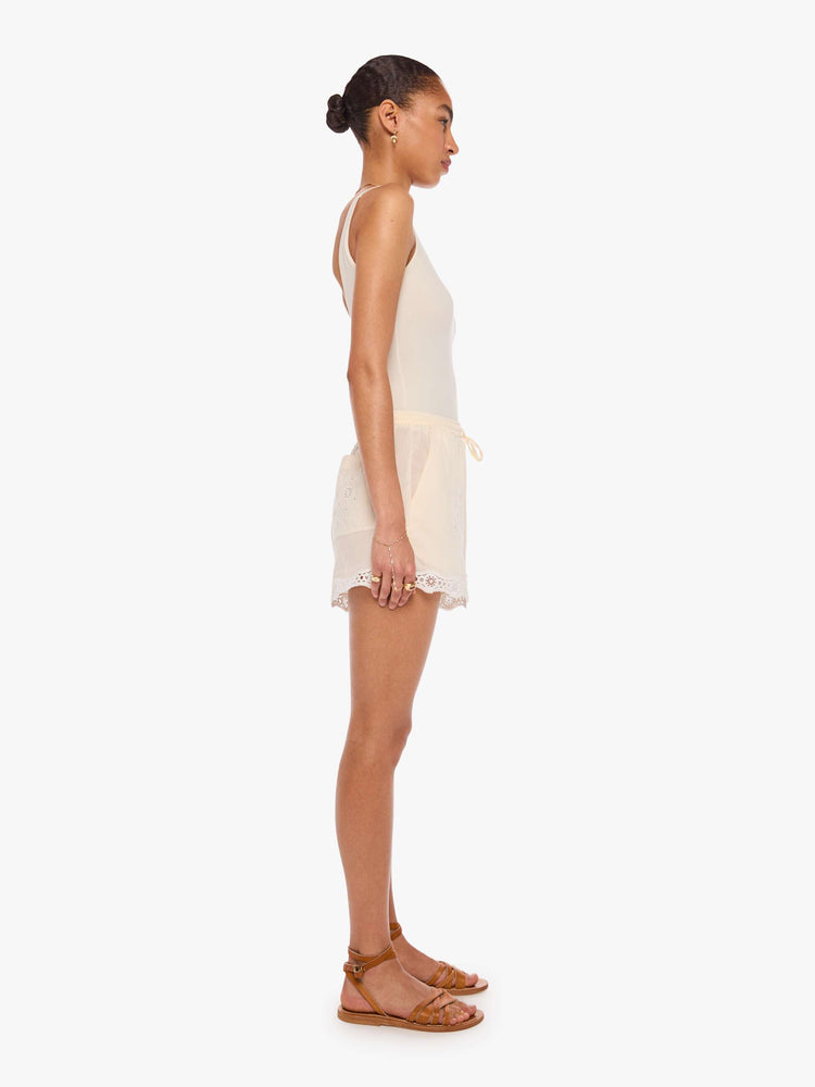 Side view of a woman in off-white short with pleats, embroidery, eyelet details and a scalloped hem.