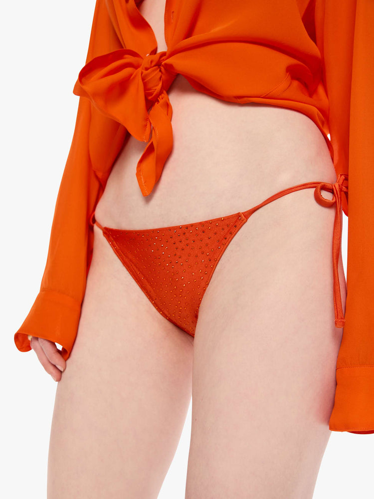Close up view of a woman in string bikini bottom from SPRWMN with decorative rhinestones throughout in orange, the bikini bottoms have a low rise and tie closures.