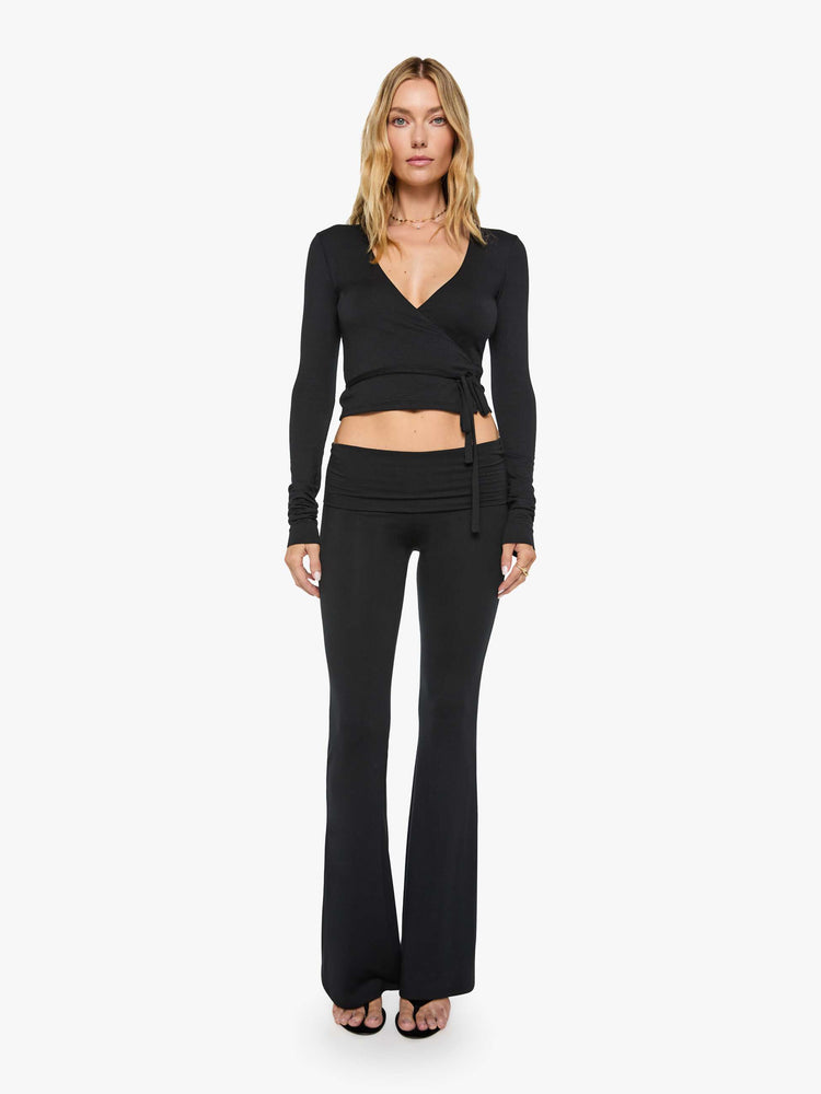 Full body view of a woman in a black top with a deep V-neck, long sleeves, a tied waist and a cropped, narrow fit.