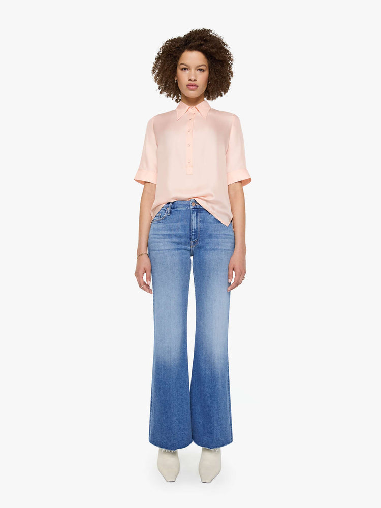 Full body view of a woman in a light rose pink shirt with a collared neckline with an extra-long button placket, elbow-length short sleeves and a loose, boxy fit.