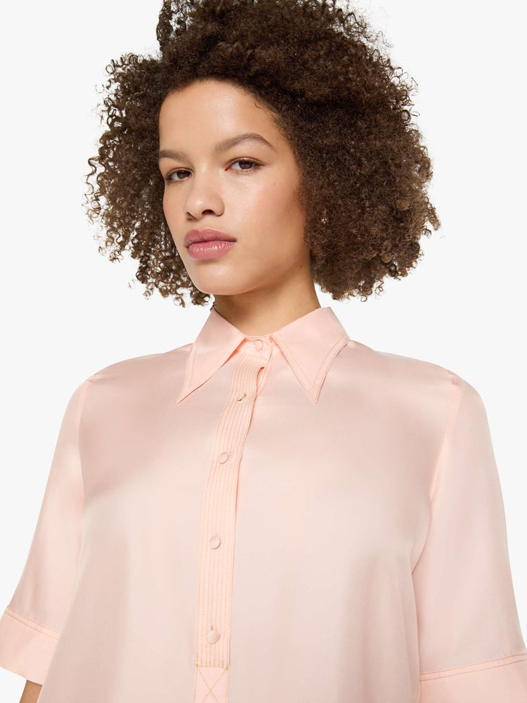 Close up view of a woman in a light rose pink shirt with a collared neckline with an extra-long button placket, elbow-length short sleeves and a loose, boxy fit.