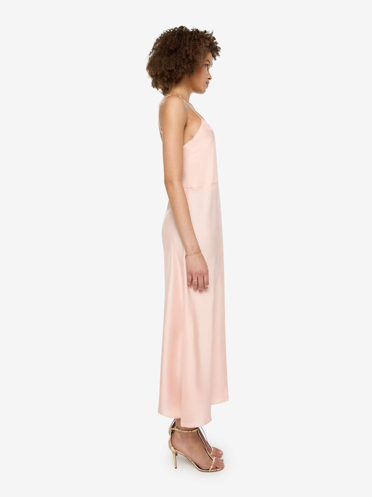 Side view of a woman in a light rose pink dress with thin spaghetti straps, a curved V-neck, a loose, flowy skirt and an ankle-length hem.