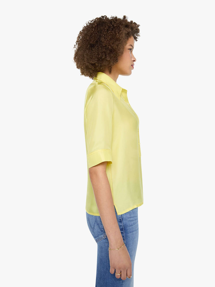 Side view of a woman in a light yellow shirt with collared neckline with an extra-long button placket, elbow-length short sleeves and a loose, boxy fit.