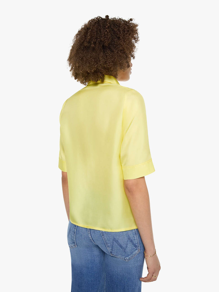 Back view of a woman in a light yellow shirt with collared neckline with an extra-long button placket, elbow-length short sleeves and a loose, boxy fit.